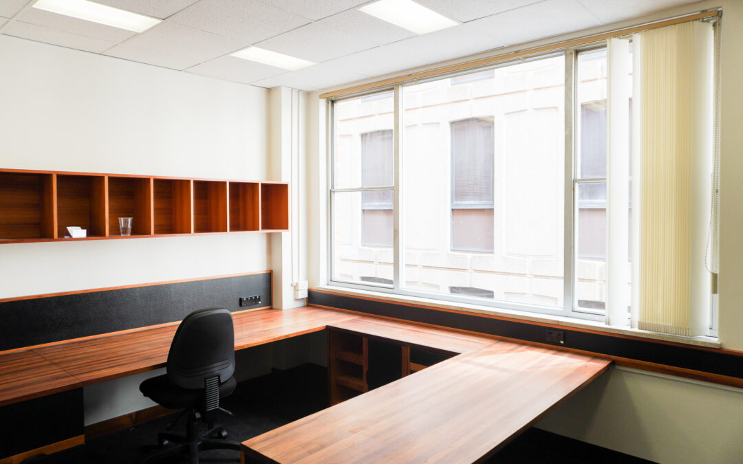 Corner office with plenty of natural light, wooden desks and storage, and a gaslift chair.