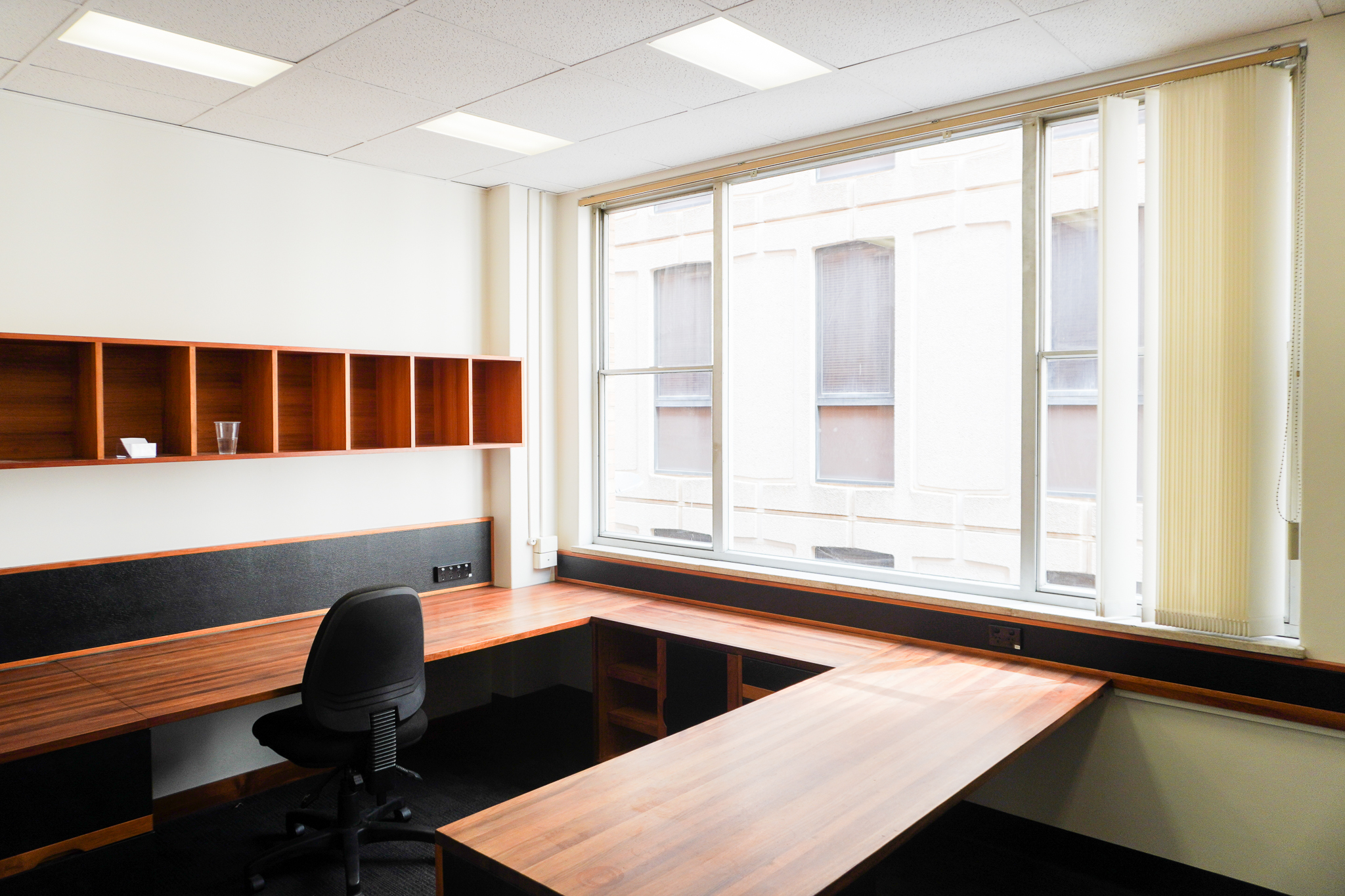 Corner office with plenty of natural light, wooden desks and storage, and a gaslift chair.