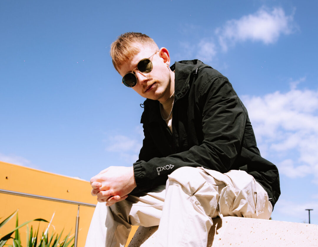 Alexander flood sitting down on a sunny day wearing sunglasses, a dark jumper and light-coloured pants.