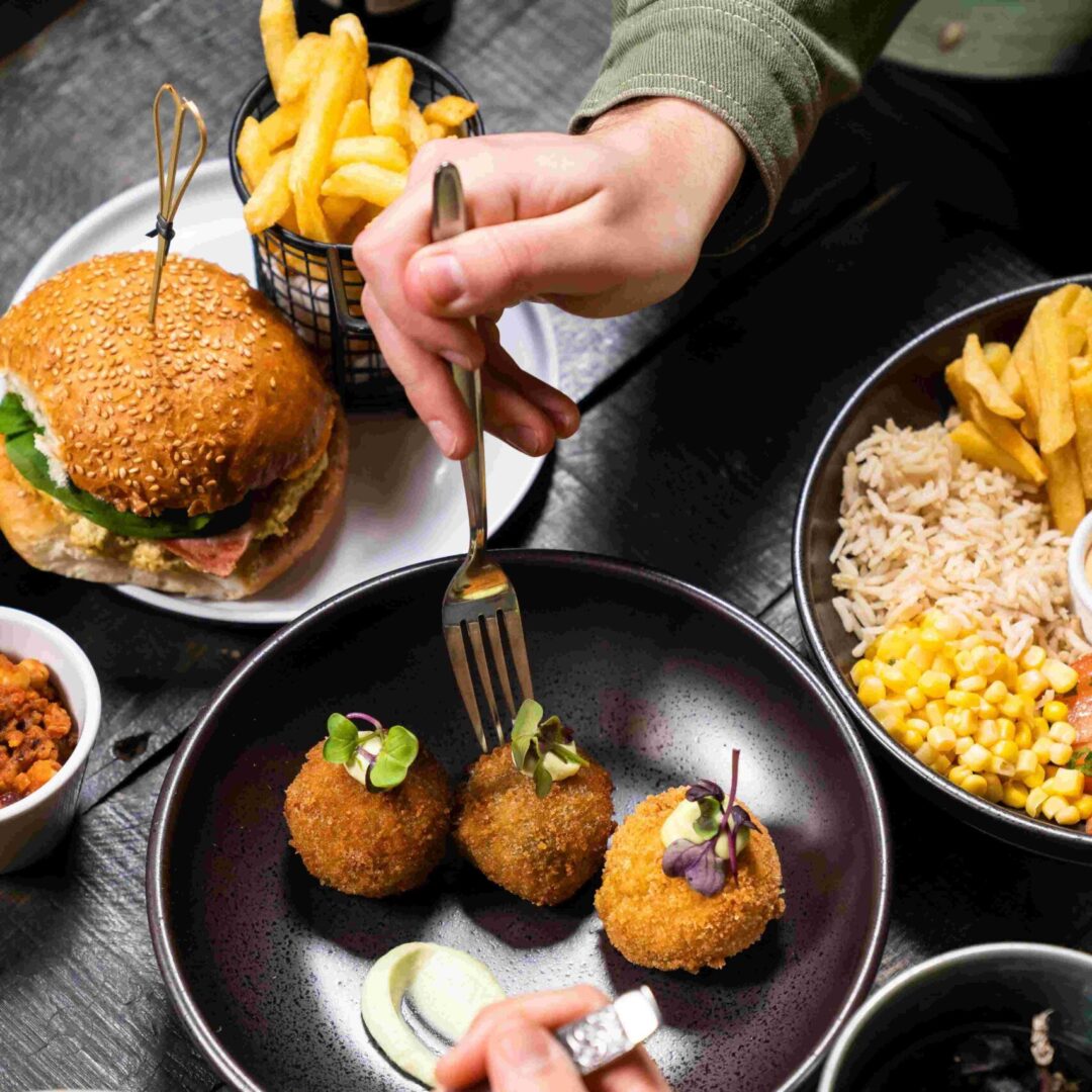 Hand using fork to scoop up arancini, with a burger and fries in the background.