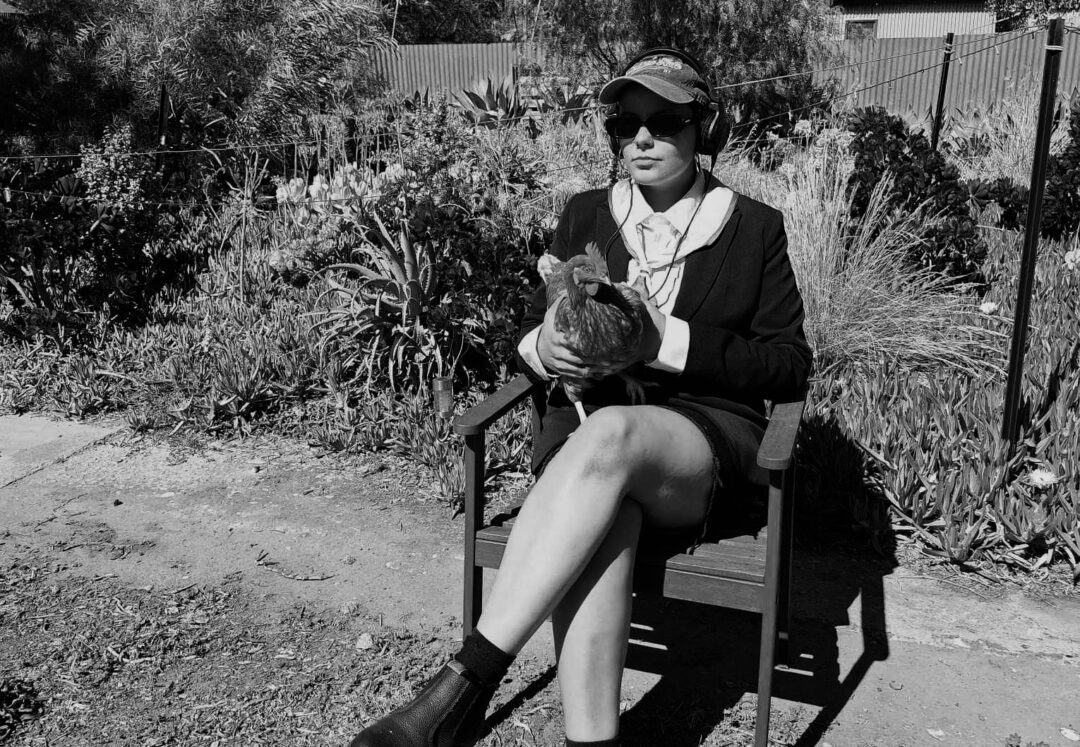 Black and white photo of Romi sitting on a chair in a backyard, wearing a cap and holding a chicken.