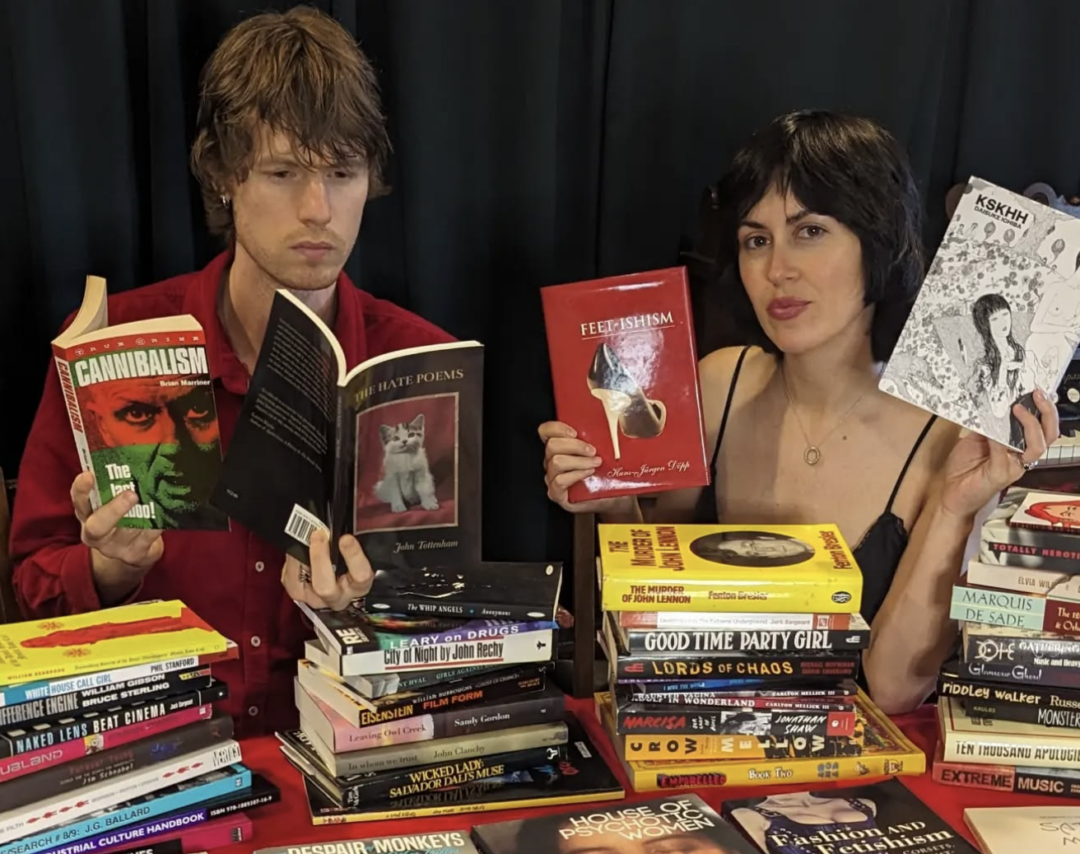 Geordie and Mel sitting at a table full of books. Geordie is reading while Mel holds up two books.