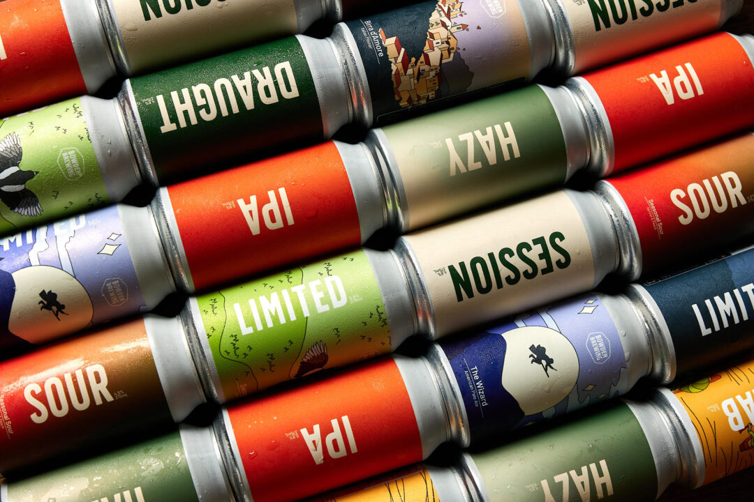 Lots of colourful beer cans stacked together at a 45 degree angle, in greens and oranges.