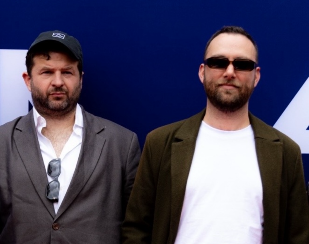 Two members of Bad Dreems, wearing suit jackets, sunglasses and baseball caps with a photo wall in the background.