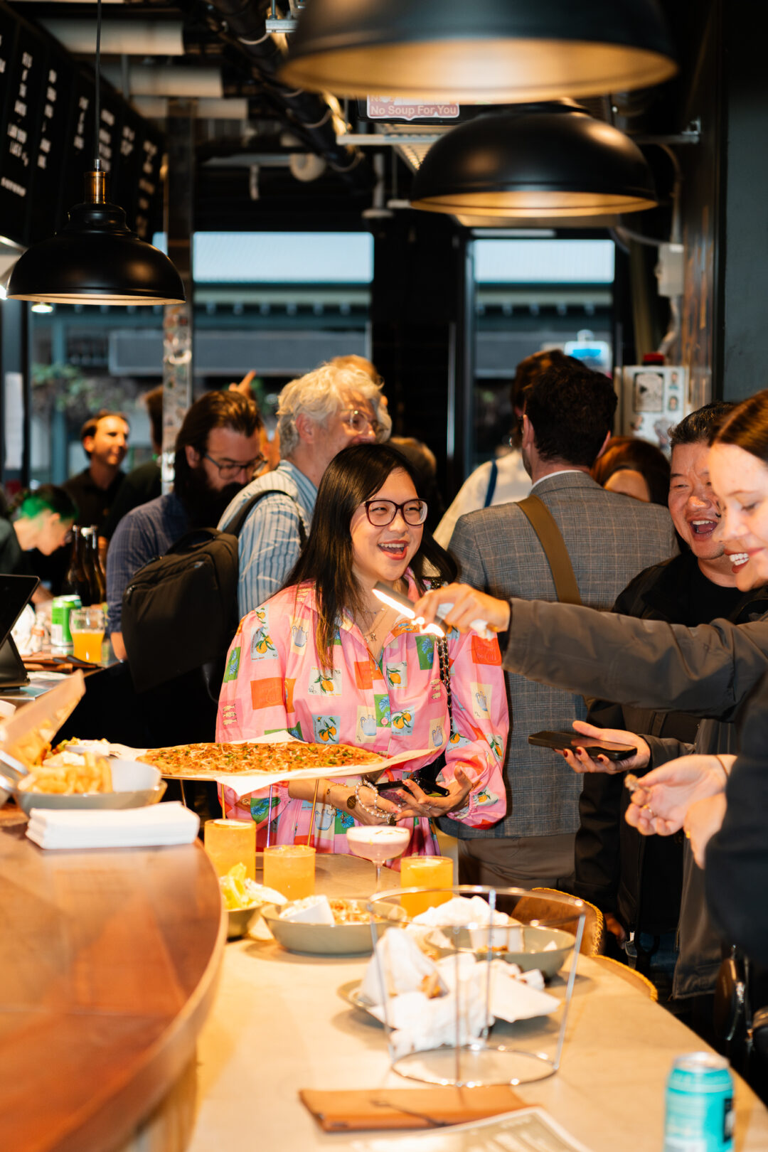 People gathered around wooden bar, sharing pizza. Woman in foreground is wearing a bright pink shirt and laughing.