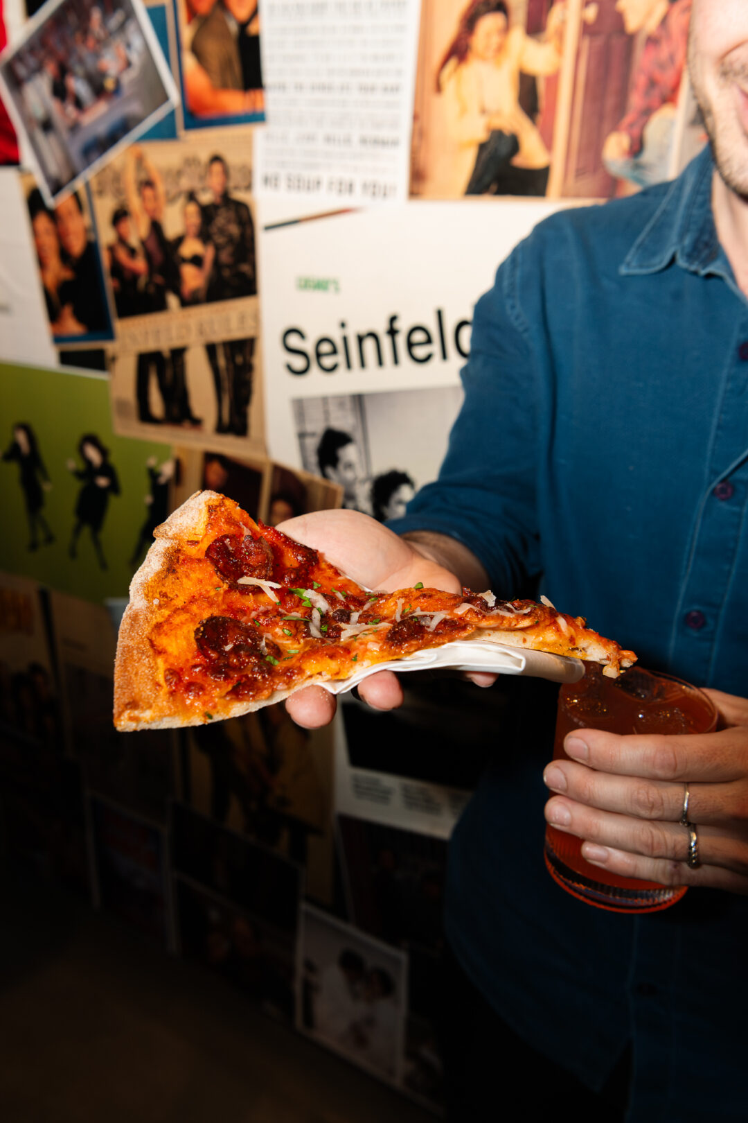 Diner wearing a blue shirt, holding a slice of pizza with posters in the background.