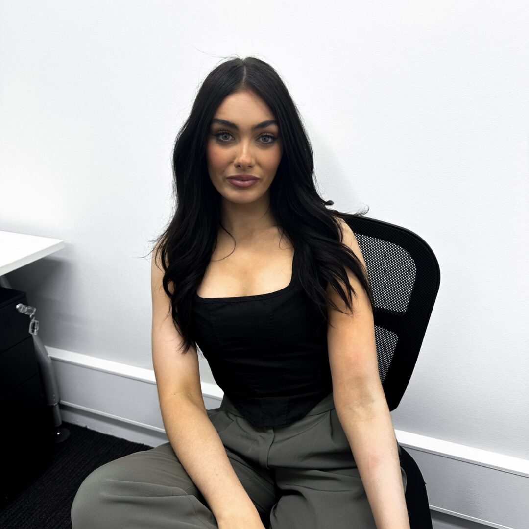 Shahna Smith sitting in a gas lift chair in her office, with a white wall. She has long dark hair and is wearing black and grey.