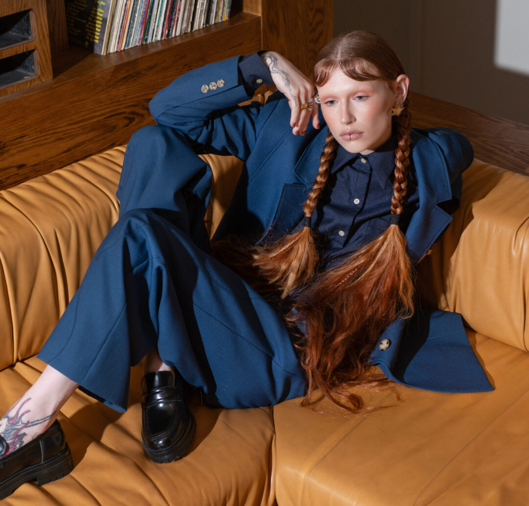 Model reclining on tan-coloured couch, wearing a slouchy blue suit. She has long red plaits.