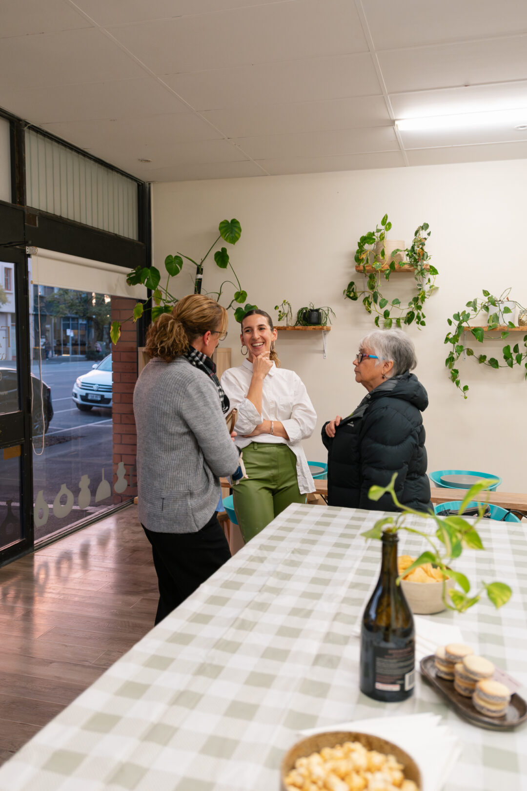 Owner Agus wearing a white shirt and green pants, laughing with two guests with plants in the background.
