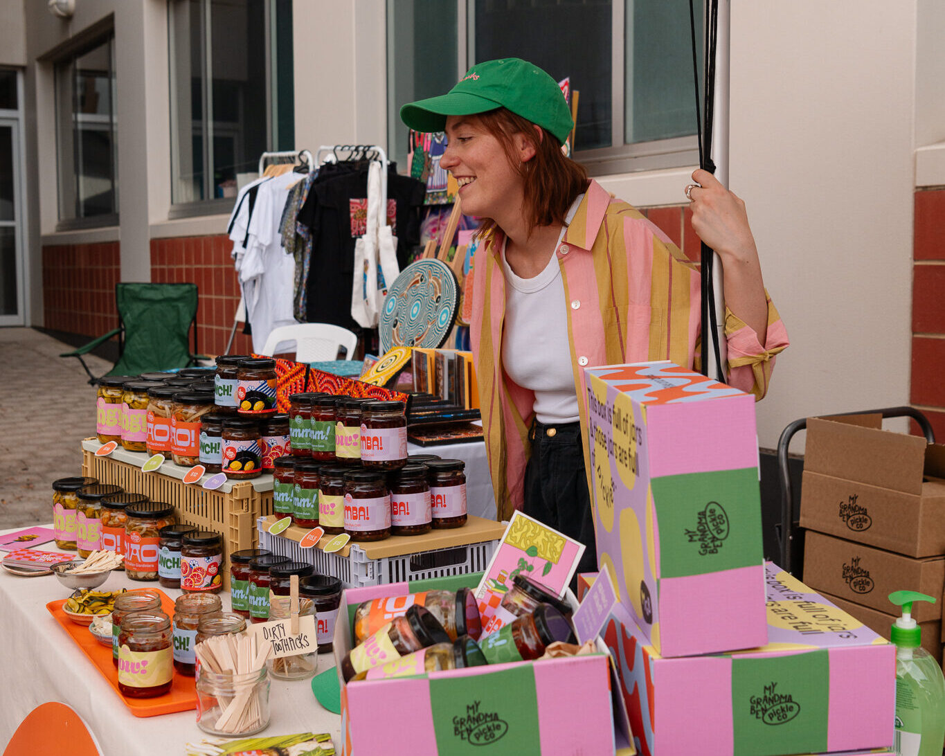 westbound-stallholder Market stall seller wearing green baseball cap and selling brightly coloured jars of product.
