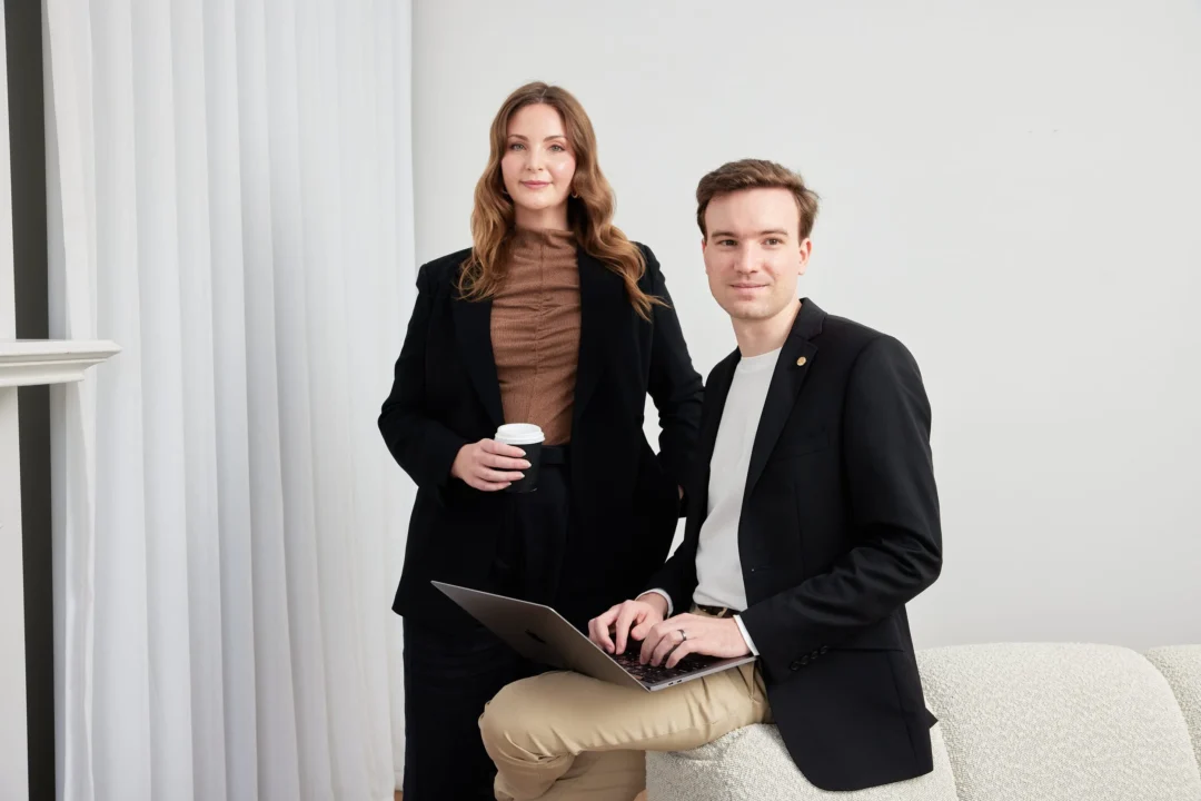 Woman standing and man leaning on couch whilst using a laptop.