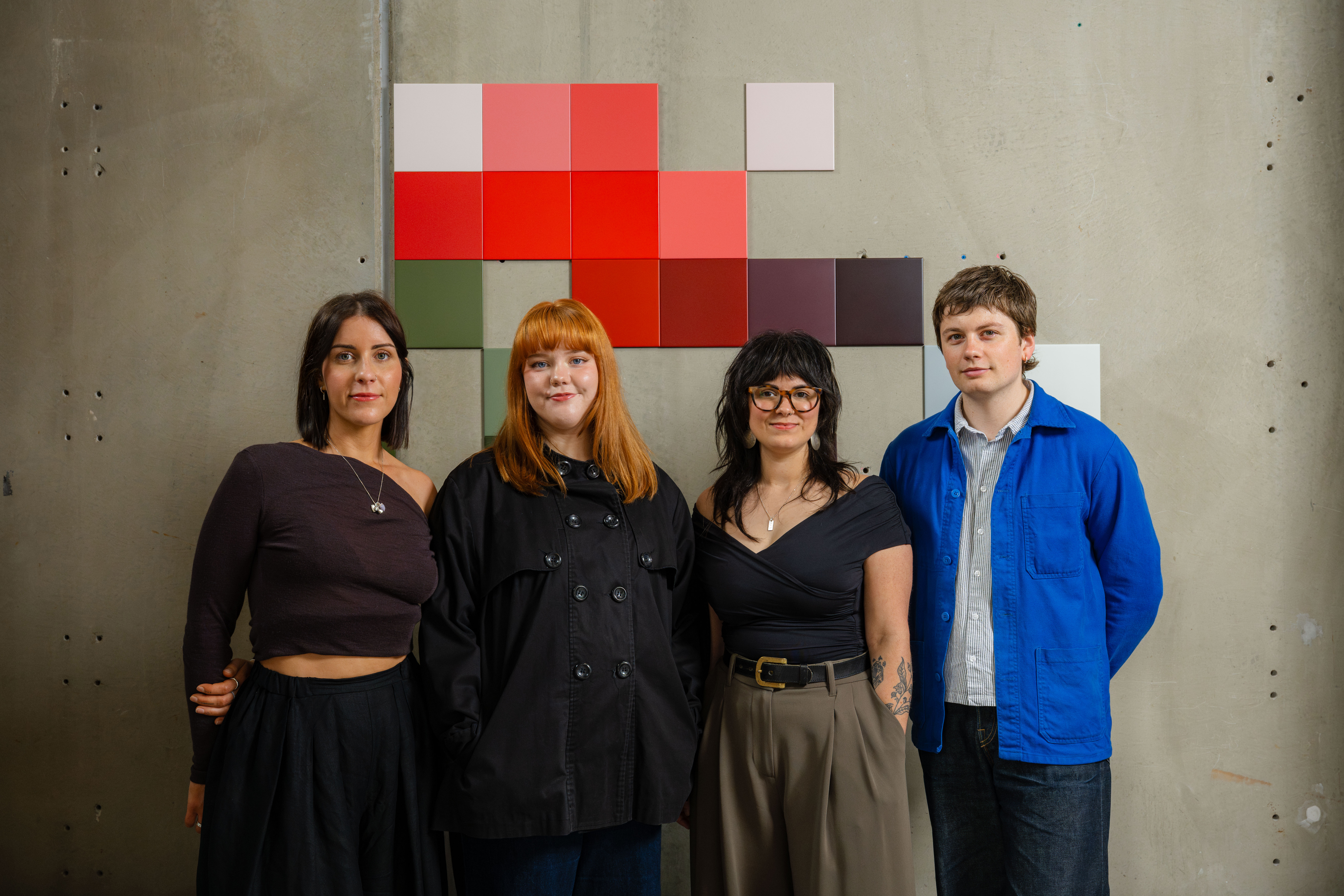 Four people standing in a row against a concrete wall with a colourful grid of square tiles behind them. They are smiling softly at the camera; three are wearing dark-toned outfits and one is wearing a bright blue jacket.