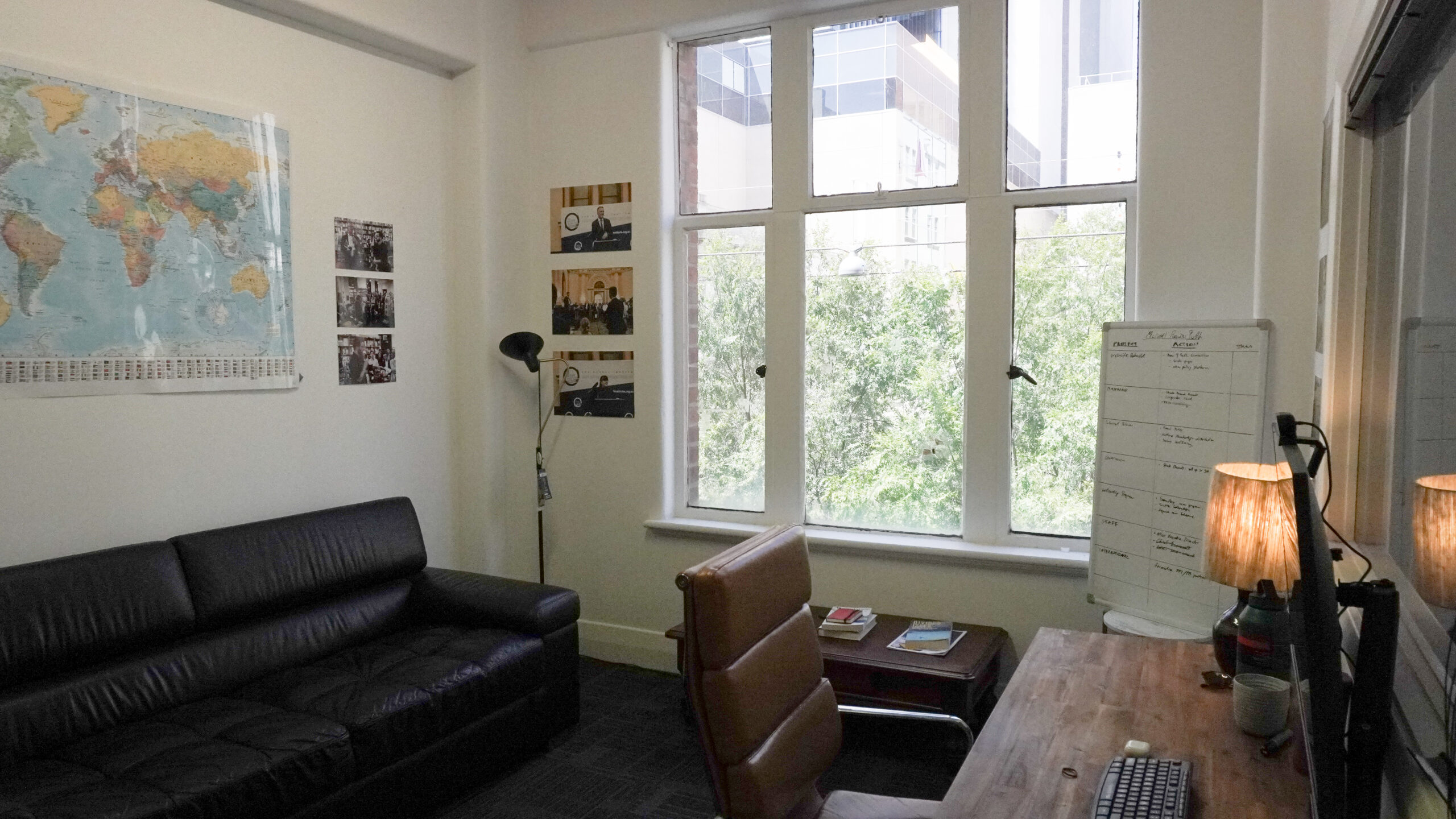 Office with white walls, large windows and lots of natural light, with a desk, couch and lamp in the foreground.