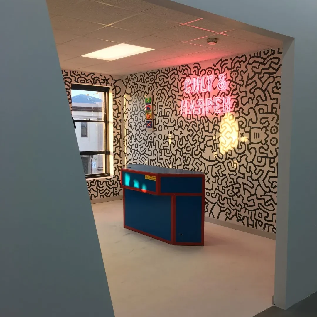 Angular view of blue reception desk, with black and white mural and pink neon sign behind it.