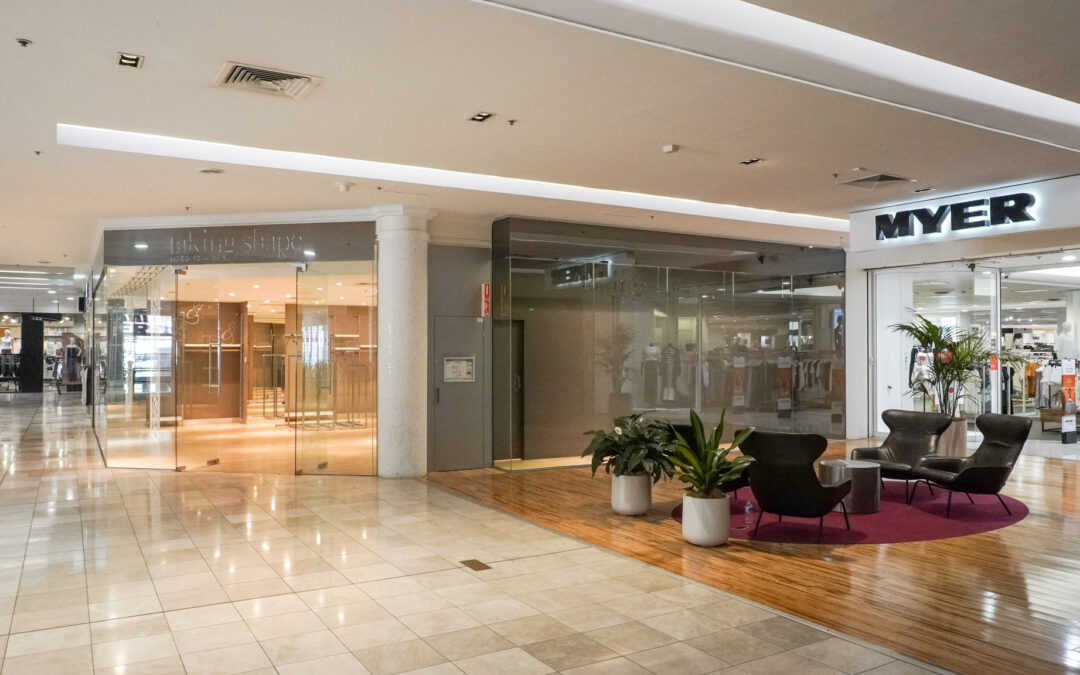 Spacious indoor shopping mall corridor with polished tile floors, featuring a Myer department store on the right, a vacant glass-front retail space on the left, and a small seating area with modern chairs and potted plants on a wooden platform.