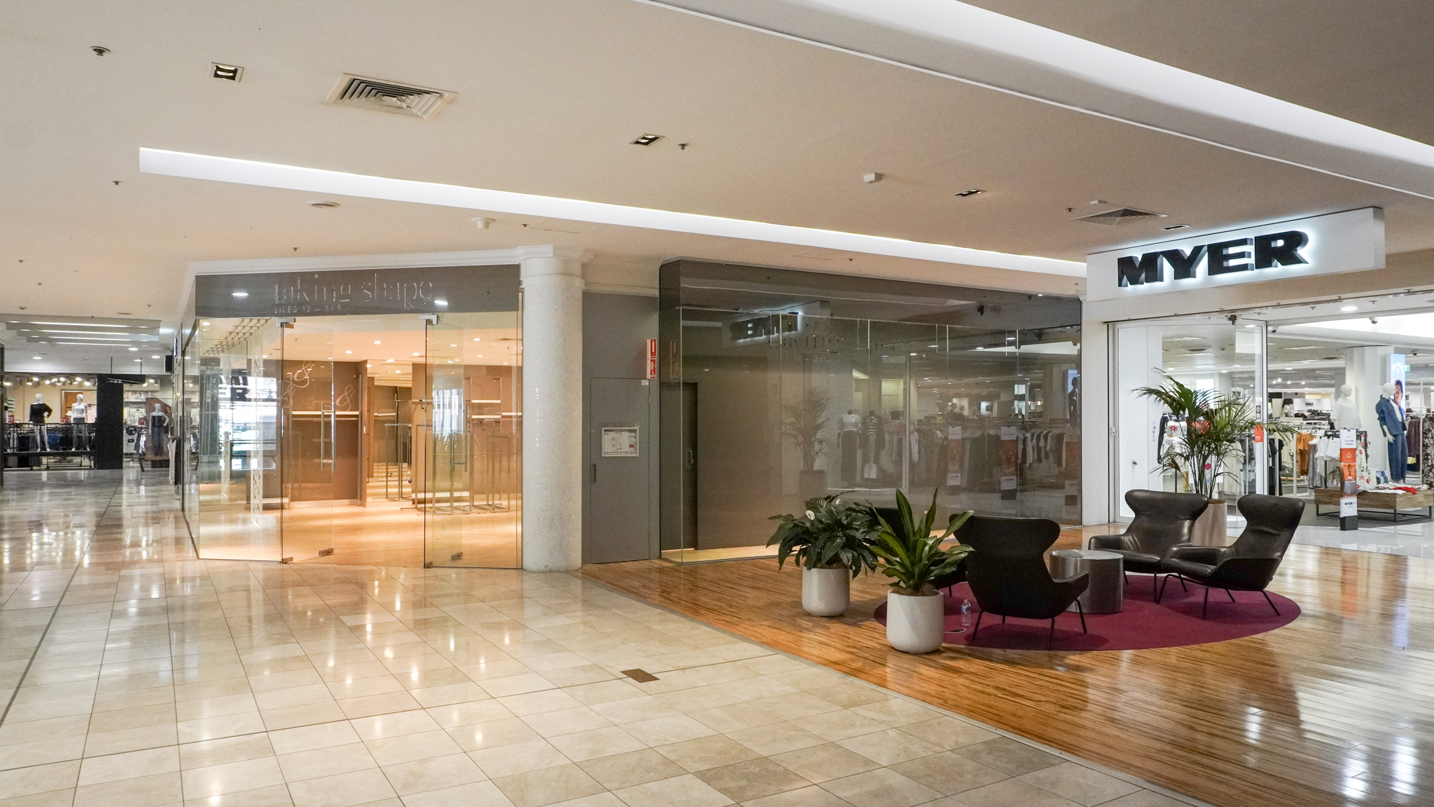 Spacious indoor shopping mall corridor with polished tile floors, featuring a Myer department store on the right, a vacant glass-front retail space on the left, and a small seating area with modern chairs and potted plants on a wooden platform.