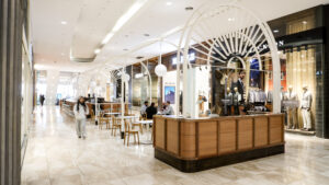 Modern mall interior with stylish wooden seating area under white archways. People are seated at tables, surrounded by clothing store displays.