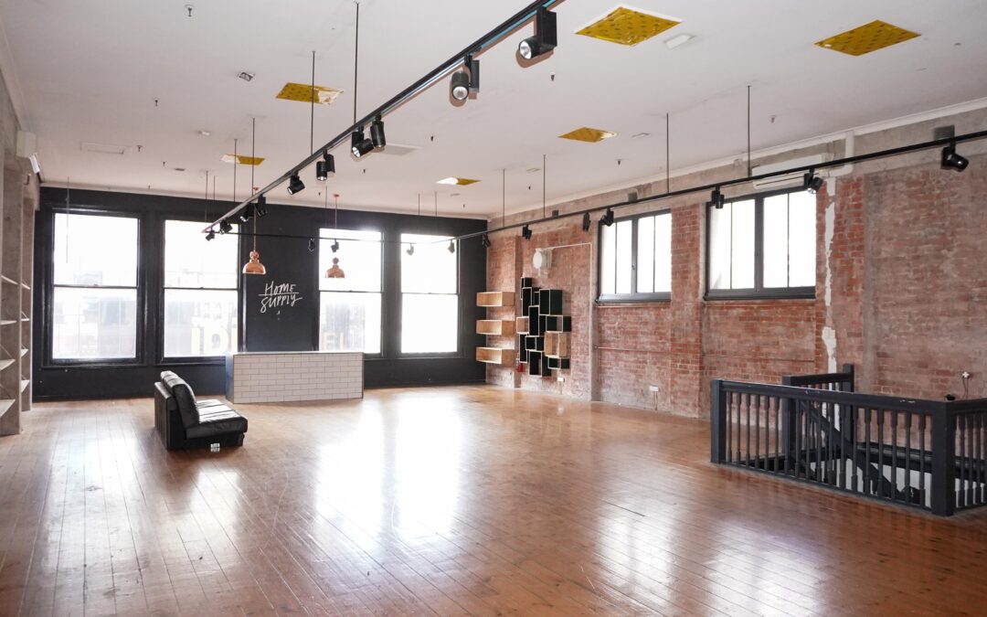 Light-filled heritage commercial space with polished timber floors, exposed red brick walls and large street-facing windows. Track lighting, high ceilings and built-in shelving frame a flexible open-plan interior, with a central reception counter and staircase leading to a lower level.