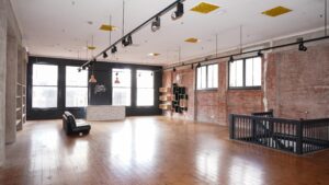 Light-filled heritage commercial space with polished timber floors, exposed red brick walls and large street-facing windows. Track lighting, high ceilings and built-in shelving frame a flexible open-plan interior, with a central reception counter and staircase leading to a lower level.