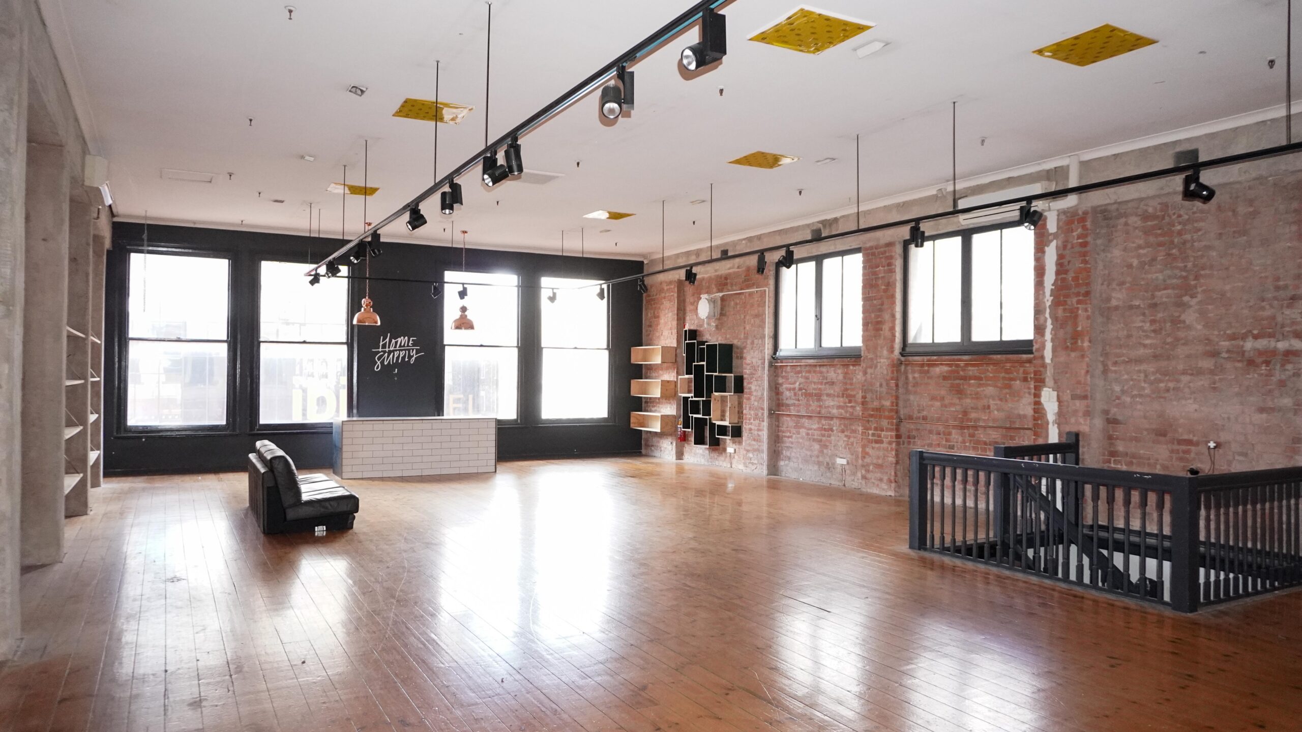 Light-filled heritage commercial space with polished timber floors, exposed red brick walls and large street-facing windows. Track lighting, high ceilings and built-in shelving frame a flexible open-plan interior, with a central reception counter and staircase leading to a lower level.