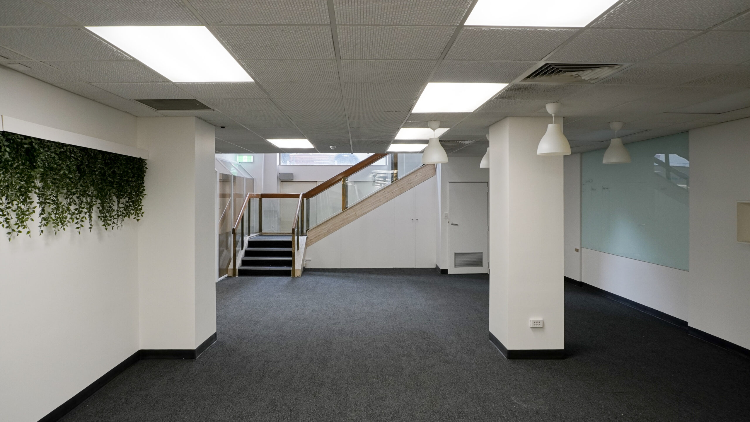 Interior of a large open-plan commercial space with carpeted floors, white walls, overhead lighting, pendant lights and a staircase with glass and timber railing.