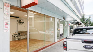 Street view of a vacant retail space with full-height glass frontage, timber-look flooring and visible sinks and hot water system inside. The shopfront is on a covered city footpath beside neighbouring businesses, with a loading zone sign and parked vehicle in the foreground.