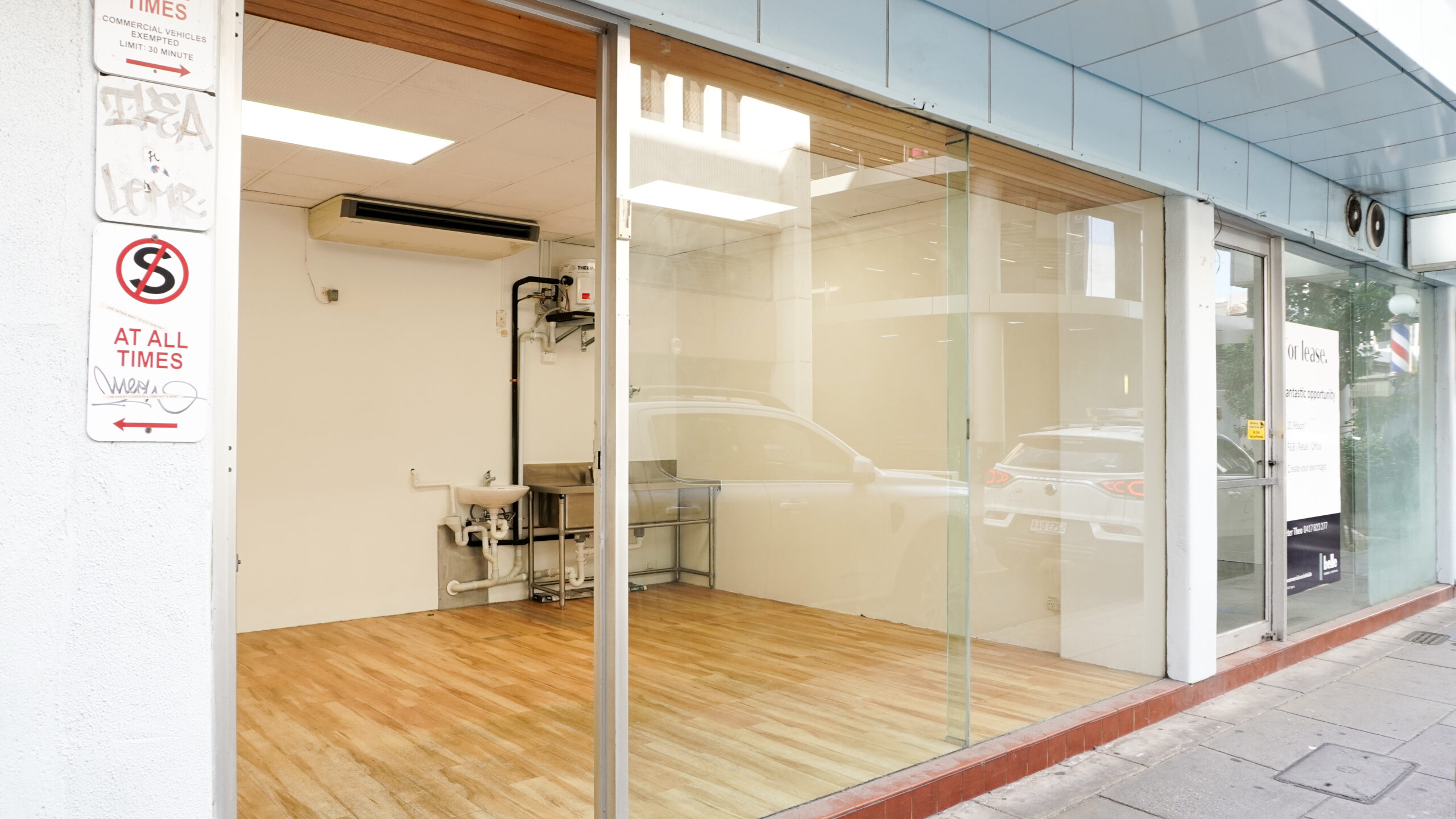 Street-facing vacant retail space with full-height glass windows, timber-look flooring, white walls, and a small stainless-steel sink and bench at the rear.