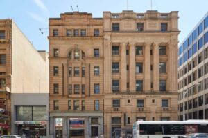 Exterior view of the historic Trustee Building at 18–22 Grenfell Street, featuring an ornate sandstone façade, large columns and classical architectural details. Modern commercial buildings and a city bus are visible along the street.